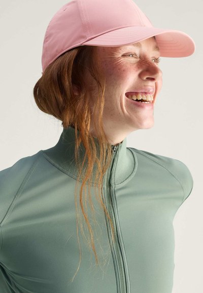 Light green zip-up top with a high collar, paired with a soft pink cap. Features smooth texture and relaxed fit, with loose hair visible.