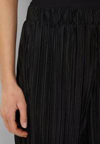Pantalons plissés noirs avec une finition texturée et brillante. Taille élastique avec des coutures verticales, main placée sur le tissu pour plus de détail.