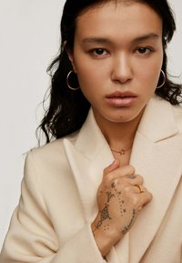 Abrigo de lana color crema con solapas de muesca, que presenta pendientes de aro dorados y un collar con estrella dorada. Mano tatuada en posición de agarre.