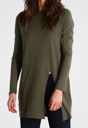 Frau trägt eine olivgrüne Tunika mit langen Ärmeln und einem vorderen Schlitz, kombiniert mit schwarzen Hosen, stehend vor einem einfarbigen Hintergrund.