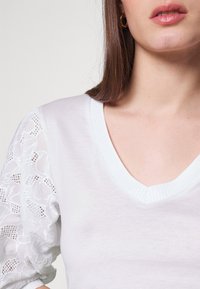 Bílé tričko s výstřihem do V a texturovanými poloprůhlednými krajkovými balónovými rukávy. Hladké bavlněné tělo s žebrovaným lemem na krku a jemným zdobením.