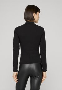Crni top dugih rukava s imitacijom polo ovratnika, ukrašen teksturiranim čipkastim detaljima na rukavima, uparen sa sjajnim crnim tajicama.