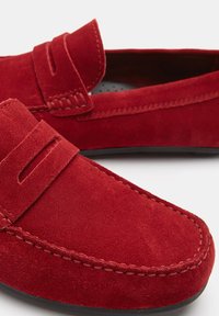 Crvene suede loafers s prošivenim detaljem preko gornjeg dijela i niskoprofilnom crnom gumennom potplatu. Glatka tekstura i minimalistički dizajn.