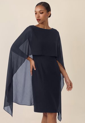 Kobieta w granatowej sukience do kolan z przezroczystymi rękawami w formie peleryny, patrząca w bok na beżowym tle.