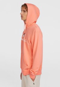Helles orange Hoodie mit einer Fronttasche und einer Kapuze mit Kordelzug. Mit einem weißen Logo auf der Brust. Glatte Textur, lockere Passform.