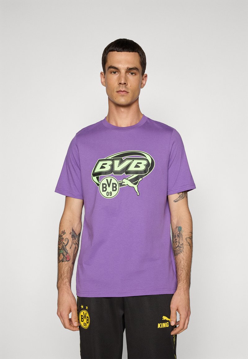 Mand iført en lila BVB-logo T-shirt og sorte shorts med gul BVB- og Puma-branding, stående mod en ensfarvet hvid baggrund.