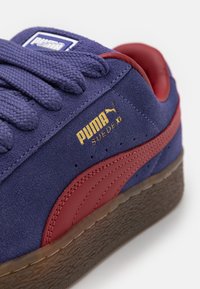 Puma suède sneakers met een marineblauwe bovenkant, rode leren accenten en een gestructureerde bruine rubberen zool. Gouden branding aan de zijkant.