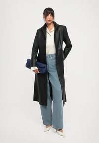 Kvinna i svart läderkappa, vit skjorta, vida jeans, vita klackar, som håller en blå texturerad klutchväska.