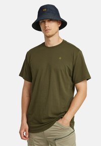 Camiseta verde oliva con cuello redondo y mangas cortas, combinada con un sombrero de cubo de denim azul oscuro, que presenta un pequeño logo amarillo.
