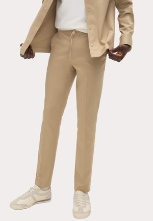 Person iført beige skræddersyede bukser med snøre, matchende beige skjorte med knapper, hvid t-shirt og hvide sneakers.