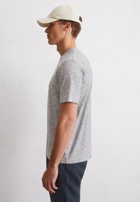 Grijs gestreept T-shirt met korte mouwen en een ronde halslijn, gecombineerd met donkere broek en een lichtgekleurde pet, vanuit de zijaanzicht bekeken.