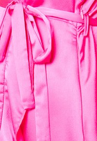 Tecido de cetim rosa brilhante com uma textura suave, apresentando um detalhe de cintura atada e elementos de design fluidos que criam uma silhueta descontraída.