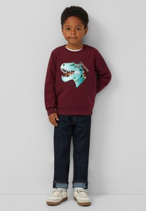 Bordeaux sweatshirt met een gestructureerde blauwe dinosaurustekening. Gecombineerd met donkerblauwe spijkerbroek en witte sneakers met grijze accenten.