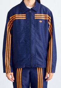 Marinjacka med orange ränder och mönstrad textur, utrustad med dragkedja framtill och sidofickor, kompletterad med matchande byxor.