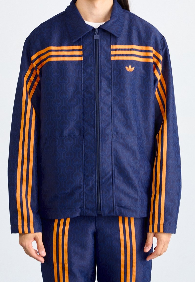 Marinjacka med orange ränder och mönstrad textur, utrustad med dragkedja framtill och sidofickor, kompletterad med matchande byxor.