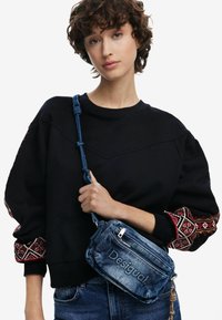 Denim-Crossbody-Tasche mit bestickten Akzenten, ausgestattet mit einem Reißverschluss und einem verstellbaren Trageriemen, kombiniert mit einem schwarzen Sweatshirt mit gemusterten Ärmeln.