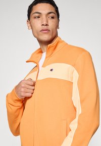 Πορτοκαλί μπουφάν με φερμουάρ, με εκρού πάνελ στο ύψος του στήθους. Διαθέτει πλαϊνές τσέπες, ριμπ μανσέτες και μικρό λογότυπο στην αριστερή πλευρά.