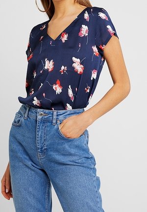 Blusa floreale blu navy con scollo a V, maniche corte e motivi di fiori rossi e bianchi sparsi, indossata con jeans blu a vita alta.
