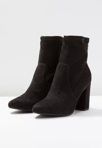 Bottines en suède noir avec un bout pointu, un talon bloc haut et un tige légèrement tombante. Texture lisse avec des détails de design minimaux.