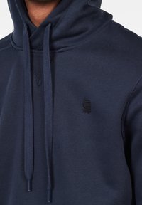 Sudadera con capucha azul marino con cordón, cuello acanalado y pequeño logo negro. Tejido suave y texturizado con un ajuste holgado.