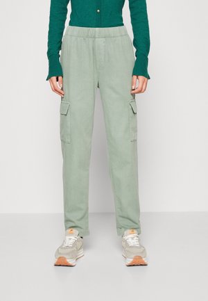Personne portant un pantalon cargo vert clair avec poches latérales, associé à des baskets beige et blanches et un haut à manches longues vert foncé.