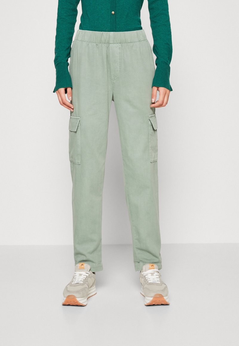 Personne portant un pantalon cargo vert clair avec poches latérales, associé à des baskets beige et blanches et un haut à manches longues vert foncé.