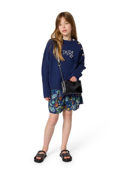 Μπλε πουλόβερ με στρογγυλή λαιμόκοψη και κουμπιά στους ώμους, συνδυασμένο με φούστα με λουλουδάτο σχέδιο, μαύρη τσάντα χιαστί και καφέ σανδάλια.