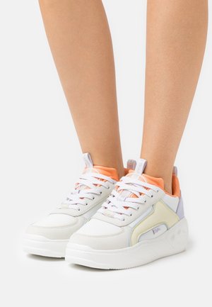 Beine, die weiße Plateau-Sneaker mit pfirsichfarbener Einfassung, lavendelfarbenem Fersenlasche und weißen Schnürsenkeln auf weißem Hintergrund tragen.