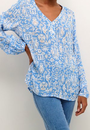 Woman wearing a blue and beige patterned long-sleeve V-neck blouse with cuffed sleeves and light blue jeans against a white background.