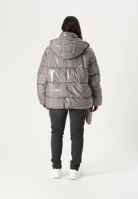 Siva puffer jakna s sjajnom završnom obradom, s horizontalnim šavovima, kapuljačom i opuštenim krojem, u kombinaciji s tamnim trapericama i bijelim tenisicama.