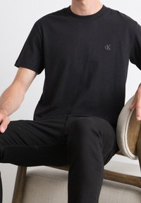 Czarny bawełniany t-shirt z krótkim rękawem, okrągłym dekoltem i małym logo na piersi. Siedzący na krześle w czarnych spodniach.