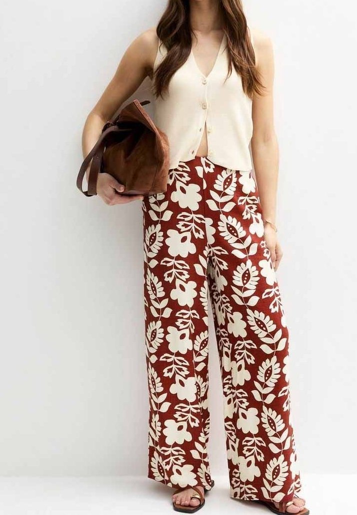 Femme portant un pantalon large couleur rouille avec imprimé floral blanc, haut crème sans manches à boutons, tenant un sac en daim marron, debout contre un mur blanc.
