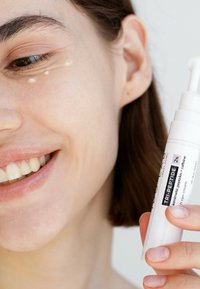 Weißer zylindrischer Tubus mit Augencreme, beschriftet "TRI-PEPTID 2%" in schwarzer Schrift. Klarer Gel wird unter dem Auge aufgetragen. Weiche Hautstruktur sichtbar.