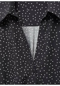 Tissu noir à pois blancs, montrant un col et une patte de boutonnage partiellement ouverte avec une doublure intérieure plus claire.