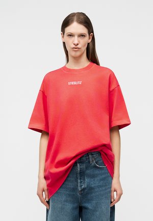 Donna con capelli lunghi indossa una T-shirt rossa oversize con la scritta "STIEGLITZ" e jeans blu su sfondo bianco.