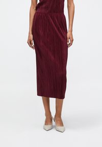 Μπορντώ πλισέ midi φούστα με κατακόρυφη υφή και ελαστική μέση, σε συνδυασμό με μπεζ μυτερές γόβες σε ουδέτερο φόντο.