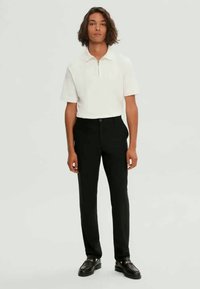 Pantalon noir sur mesure associé à un polo blanc à manches courtes avec un col zippé. Le mannequin se tient devant un fond neutre.