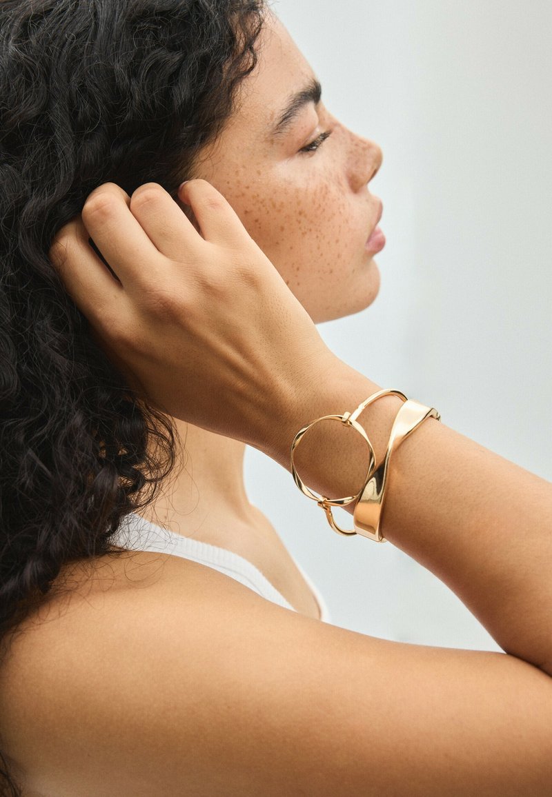 Femme aux cheveux bouclés et foncés portant deux bracelets en or, touchant ses cheveux, tournée de profil sur un fond clair uni.