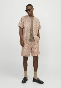 Chemise à manches courtes à motifs floraux et short assorti en beige avec des motifs rouges. T-shirt marron à col rond en dessous. Associé avec des mocassins noirs.