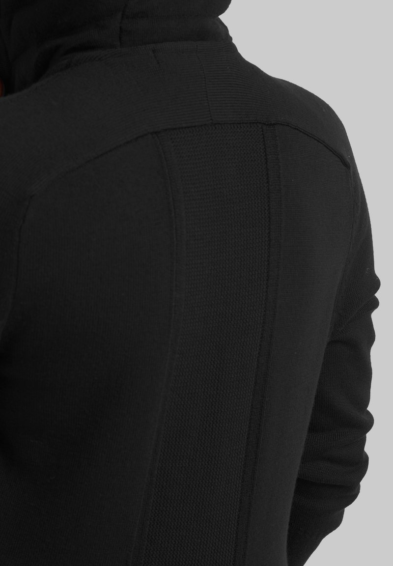 Rückansicht einer Person, die einen schwarz strukturierten Strickpullover mit gerippten Schultern und hohem Kragen vor einem grauen Hintergrund trägt.