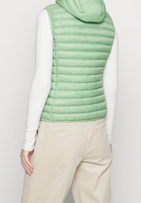 Jasnozielona, pikowana kamizelka z kapturem, skrojona w dopasowany fason, noszona na białej bluzce z długim rękawem i beżowych spodniach z korduroy.