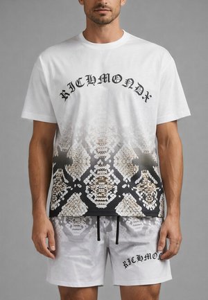 Uomo con maglietta bianca Richmond con motivo a pelle di serpente nero e beige e pantaloncini bianchi abbinati con scritta Richmond.