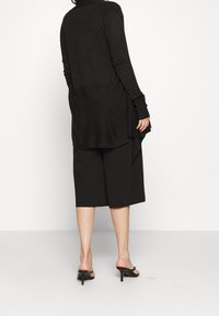 Černý top s dlouhým rukávem a volně splývající zadní částí, doplněný černými širokými šortkami. Oblečení má matnou texturu a neformální siluetu.