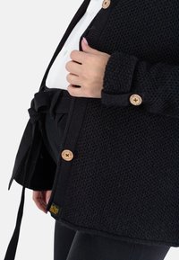 Schwarze Strickjacke mit Holzknöpfen, einer strukturierten Stoffoberfläche und einem gebundenen Taillenbund, mit einem Logo-Tag, das am Saum angenäht ist.