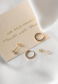 Die goldenen Ohrringe bestehen aus einem kleinen Stecker mit einem klaren Stein und einem Hoop-Design, präsentiert auf einer beigen Karte mit goldfarbigem Text. Weicher weißer Stoffhintergrund.