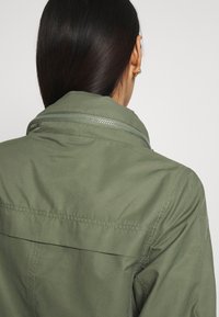 Olivengrøn jakke med høj krave, synlige syninger og lynlås. Stoffet virker let med en glat tekstur.