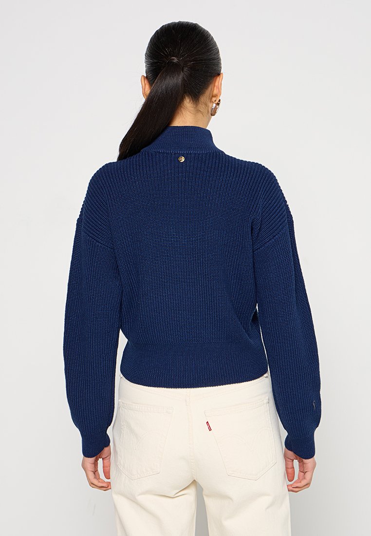 Maglione lavorato a maglia blu navy con un colletto alto, texture a costine e un piccolo dettaglio con bottone dorato sul retro. Vestibilità rilassata e spalle scese.