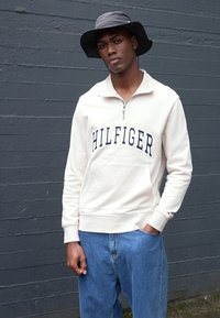 Felpa zip beige chiaro con "HILFIGER" stampato in blu navy. Abbinata a un pantalone di jeans blu. Modello che indossa un cappello a secchiello grigio.