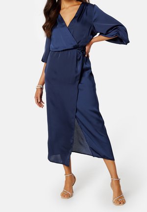 Kobieta ubrana w granatową sukienkę kopertową z satyny z rękawami do połowy przedramienia i bocznym rozcięciem, w połączeniu z sandałami na wysokim obcasie z paskami.
