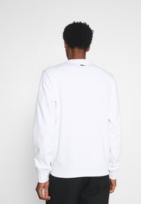 Sudadera blanca de manga larga, cuello redondo, con puños y dobladillo acanalados, que presenta un pequeño logo verde en la parte posterior, cerca del cuello.